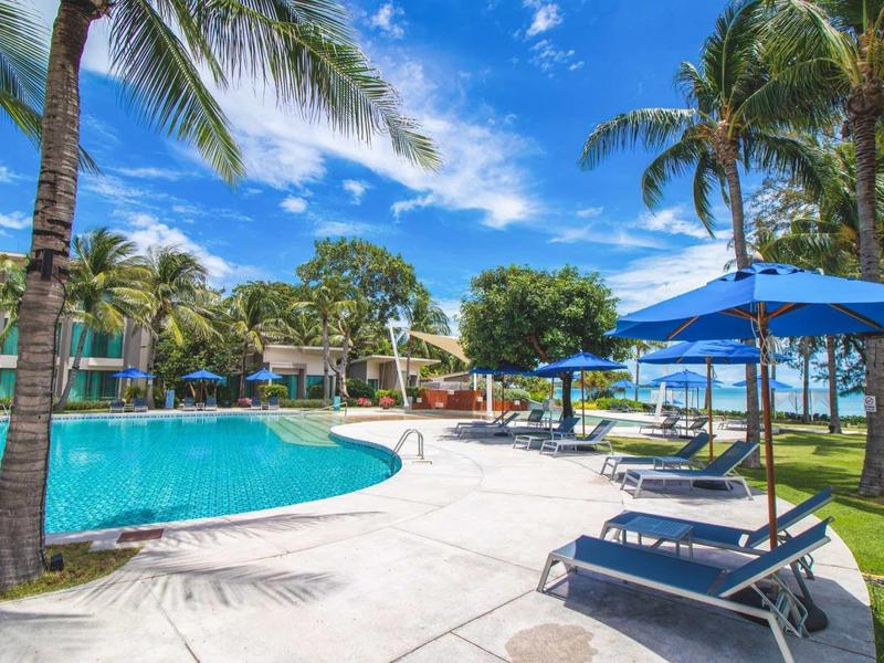 Hotel pool with lounge chairs, blue umbrellas, and palm trees under clear sky.