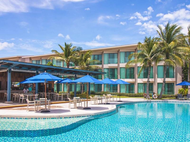 Hotel with large pool, blue umbrellas, and palm trees under a sunny sky.