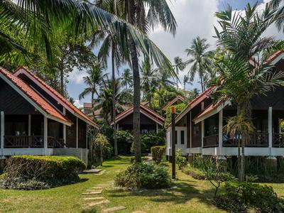 Kilka bungalowów z werandami otoczonych palmami i trawnikiem w tropikalnym otoczeniu.