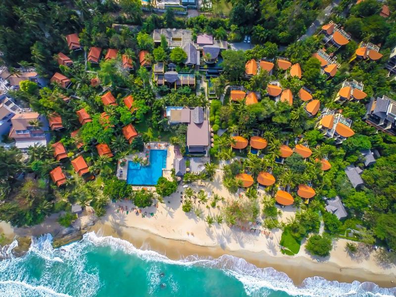 Letecký pohled na resort s červenými střechami, bazénem a pláží u tyrkysového moře.