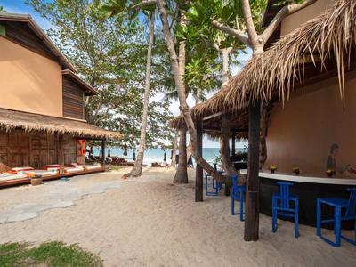 Sandiger Pfad zwischen zwei Strohdachhäusern mit Sitzgelegenheiten und Bäumen, Blick aufs Meer.