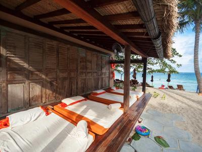 Strandbungalow mit weißen Liegen und orangen Handtüchern, Palmen und Meer im Hintergrund.