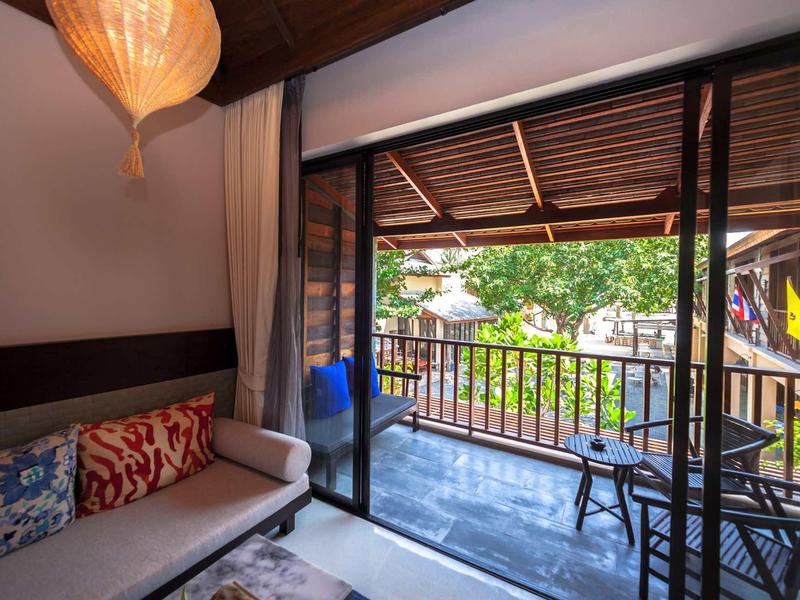 Wohnzimmer mit Sofa und Kissen, gläserne Schiebetür führt zu Balkon mit Holzgeländer und Stühlen.