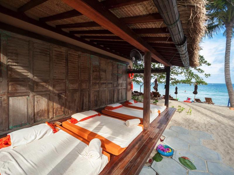 Strandbungalow mit weißen Liegen und orangen Handtüchern, Palmen und Meer im Hintergrund.