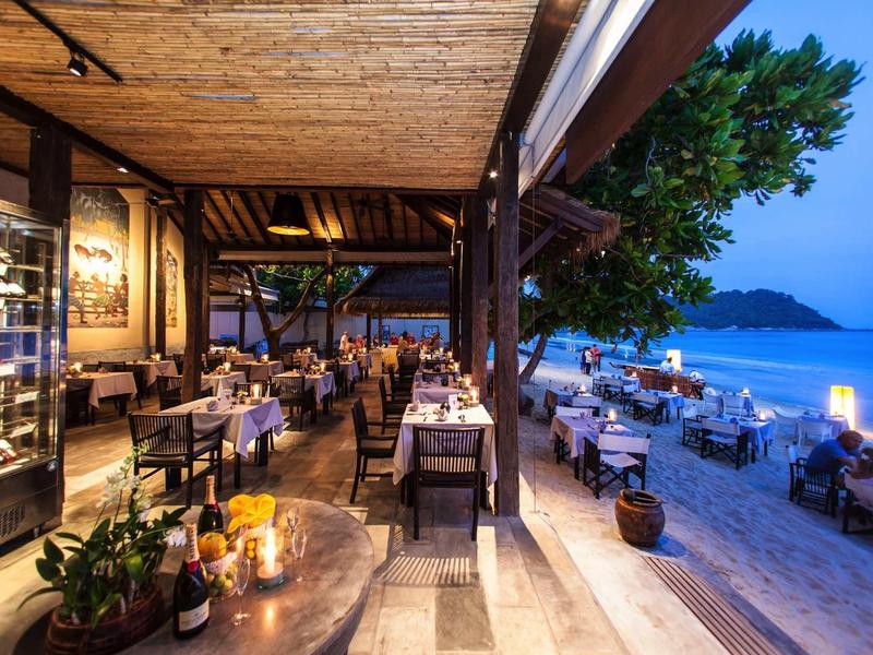 Restaurant mit gedeckten Tischen am Strand, Abendstimmung, Meerblick und dekorativer Beleuchtung.