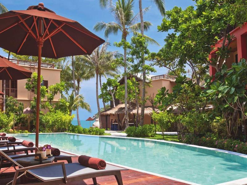 Schwimmbad mit braunen Liegestühlen und Sonnenschirmen, umgeben von grünen Pflanzen und blauen Himmel.