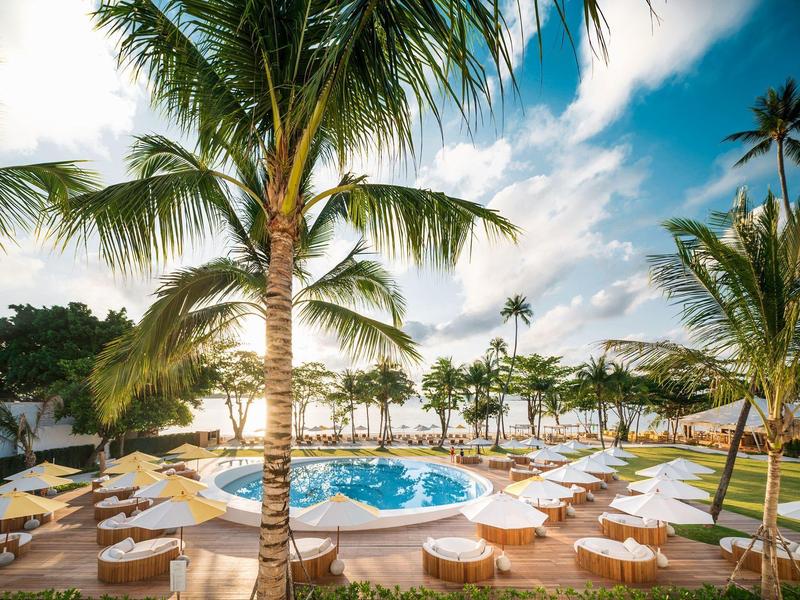 Area piscina con palme, lettini e ombrelloni sotto un cielo azzurro in un resort.