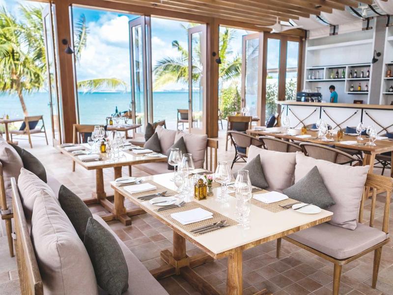 Helles Restaurant mit Holzdecke, großen Fenstern, Blick auf Palmen und Meer, gedeckten Tischen und gemütlichen Polstern.
