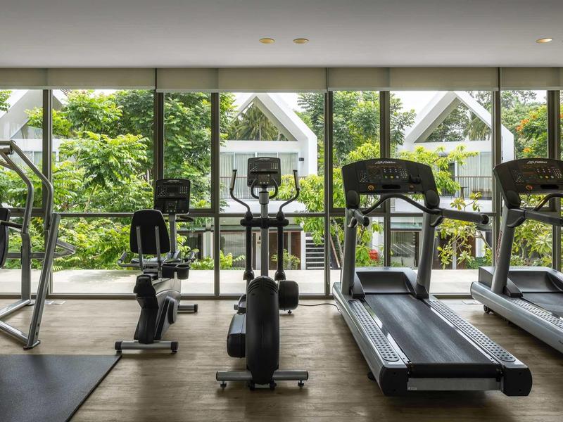 Moderner Fitnessraum mit Laufbändern und Fitnessgeräten vor großen Fenstern mit Blick auf grüne Bäume.