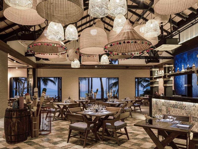 Gemütliches Restaurant mit Holztischen, runden Lampen und großen Fenstern mit Meerblick.