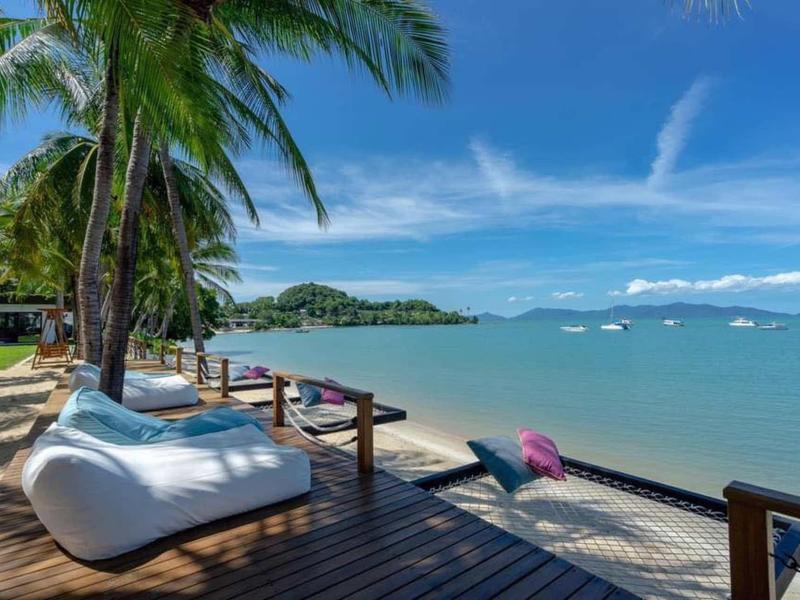 Holzterrasse mit weißen Sitzkissen und Palmen am ruhigen Meer unter blauem Himmel.
