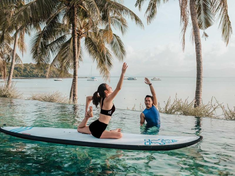 Zwei Personen sitzen auf einem Stand-up-Paddleboard in einem Pool mit Palmen im Hintergrund.