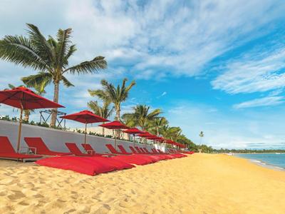 Strand mit roten Liegestühlen und Sonnenschirmen neben Palmen und blauem Himmel.
