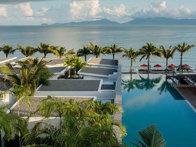 Moderner Poolbereich mit Palmen, roten Sonnenschirmen und Meerblick bei bewölktem Himmel.
