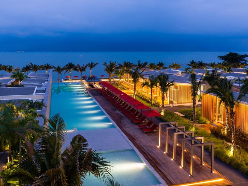 Luxuriöser Poolbereich mit Palmen, beleuchteten Wegen und Blick auf das ruhige blaue Meer bei Abenddämmerung.