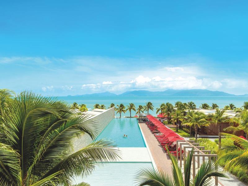 Blick auf einen Pool mit Palmen, roter Sonnenschirme und grüner Rasenfläche unter blauem Himmel.