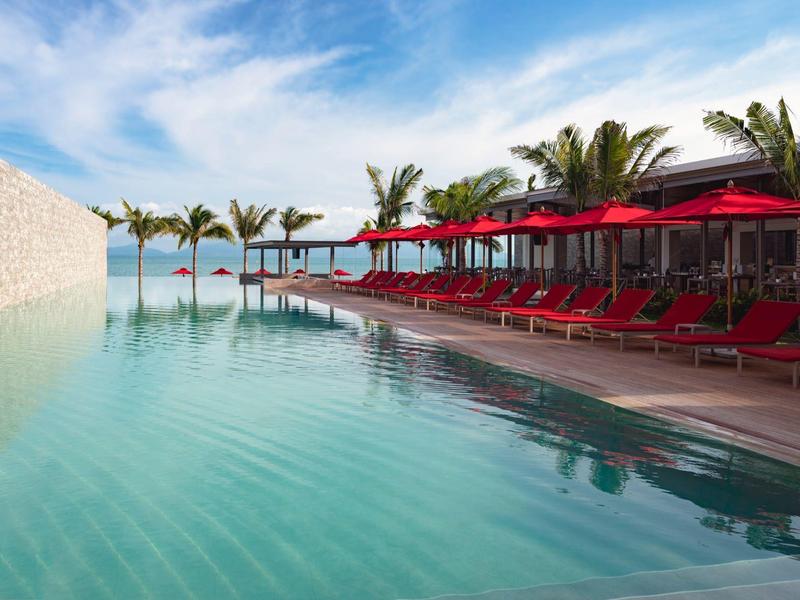 Großer Pool mit roten Liegestühlen und Palmen unter blauem Himmel am Meer.