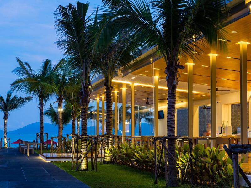 Abendlicher Blick auf eine beleuchtete Strandpromenade mit Palmen und Restaurant.