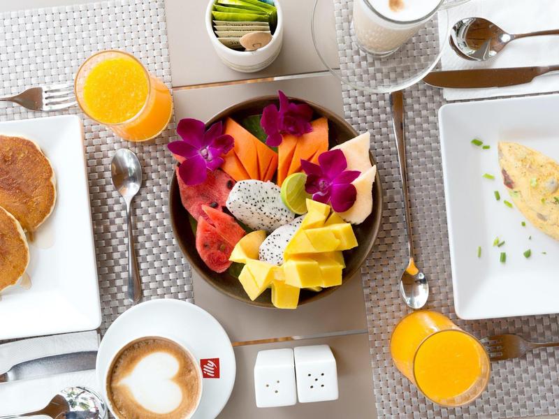 Frühstückstisch mit Teller voller frischem Obst, Pancakes, Omelett und Getränken, heller Hintergrund.