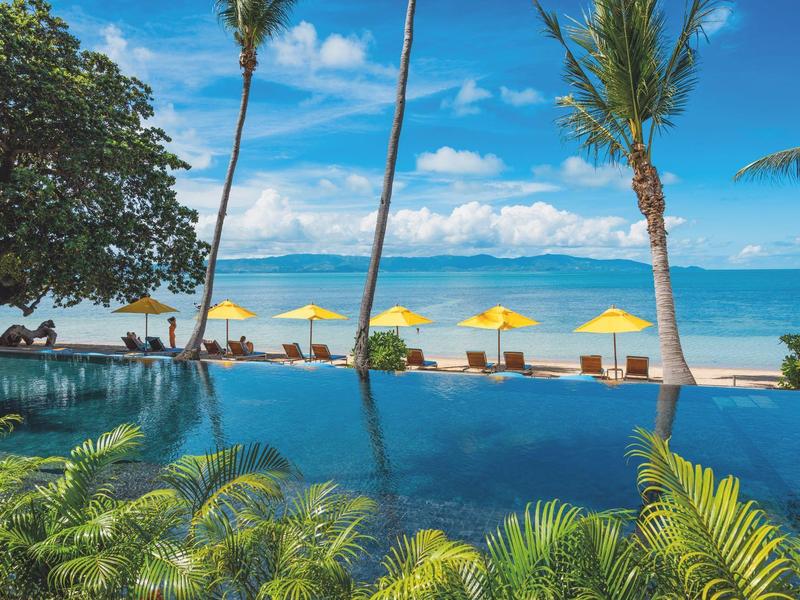 Piscina dell'hotel con vista sulla spiaggia, ombrelloni gialli e palme sotto un cielo azzurro.