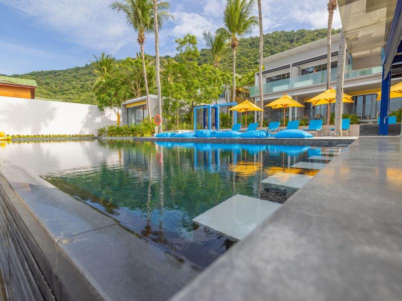 Piscina moderna dell'hotel con lettini blu e ombrelloni gialli in ambiente tropicale.