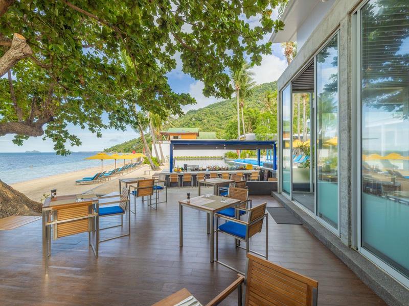 Terrazza di un bar sulla spiaggia con tavoli, sedie e vista sulla sabbia e sul mare.