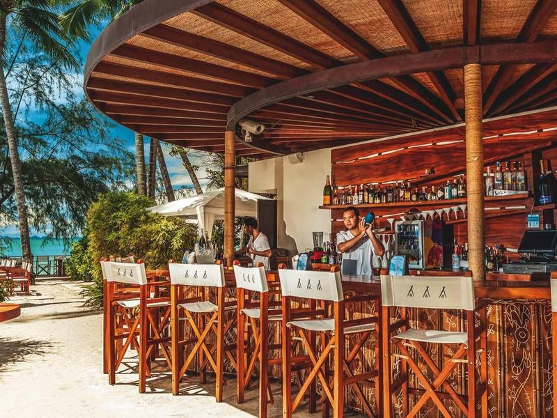 Buitenbar met houten stoelen op een strand onder palmbomen bij zonnig weer.