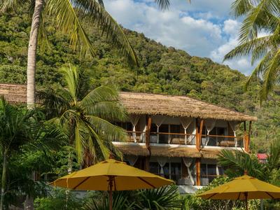 Traditionelles Gebäude mit Strohdach, Palmen und gelben Sonnenschirmen in tropischer Umgebung.