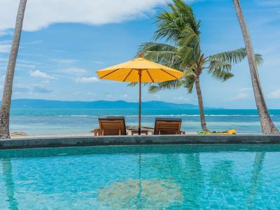 Ein Pool mit klarem Wasser vor dem Strand, zwei Liegestühle und ein gelber Sonnenschirm, Palmen im Hintergrund.