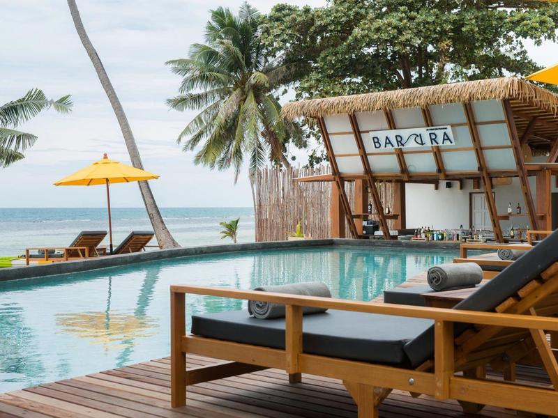 Pool mit Liegestühlen, Palmen und gelben Sonnenschirmen am Strand bei bewölktem Himmel.