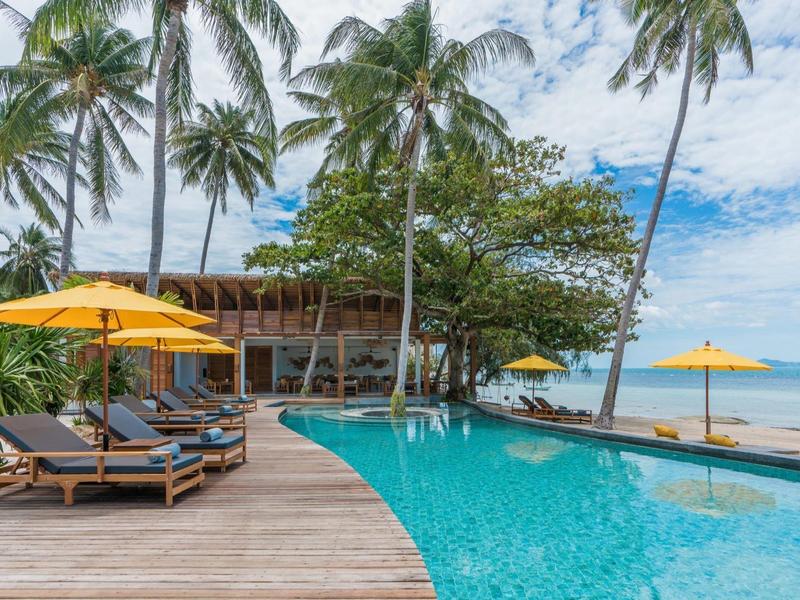 Pool mit Sonnenschirmen und Liegen, umgeben von Palmen am Strand unter blauem Himmel.