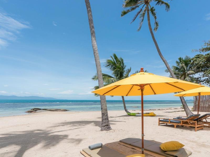 Strand mit weißem Sand, zwei Liegestühlen, gelbem Sonnenschirm und Palmen vor blauem Himmel.