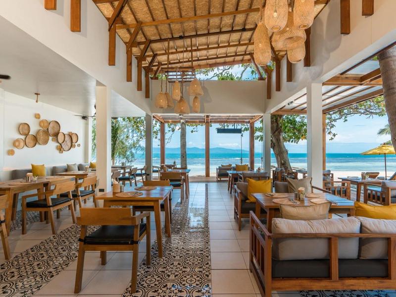 Offenes Restaurant mit Holzmöbeln, geflochtenen Lampen und Meerblick bei klarem Himmel.