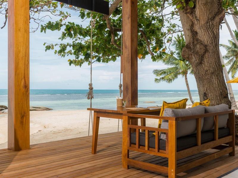 Holzterrasse mit Schaukel, Sofa und Blick auf weißen Sandstrand und ruhiges blaues Meer.