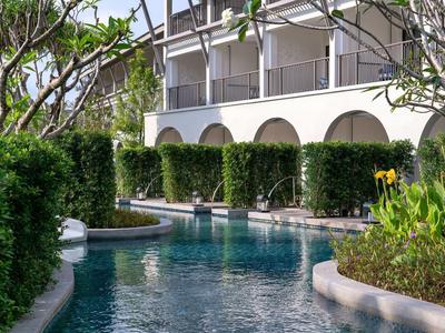 Bazén obklopený bujnou vegetací a bílou fasádou hotelu s oblouky.