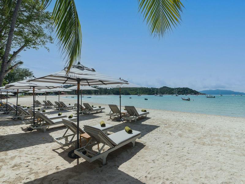 Bílá písečná pláž s lehátky a slunečníky, klidné tyrkysové moře pod modrou oblohou.