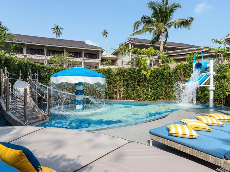 Hotelový bazén s tobogánem, modrým slunečníkem a lehátky u palem.