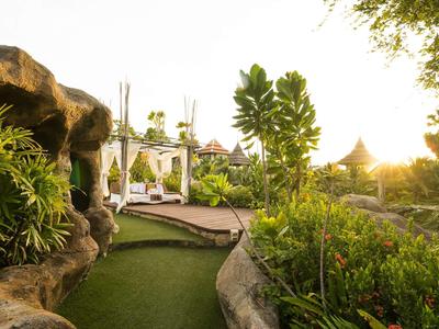 Grüner Garten mit Liegen, Pavillons und Felsen bei Sonnenuntergang in einer Hotelanlage.