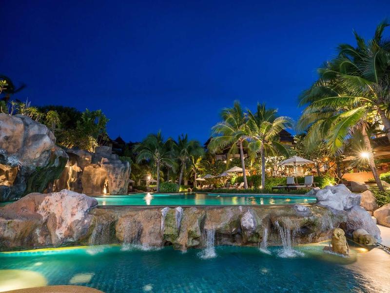 Vue nocturne d'une piscine de complexe tropical avec cascades et palmiers illuminés.