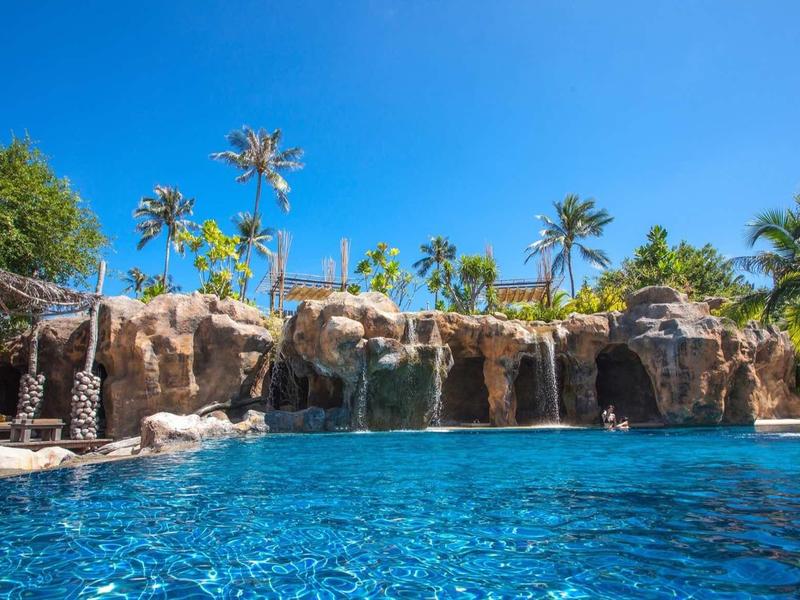 Grand bassin avec cascade, entouré de palmiers et ciel bleu dans une station hôtelière.