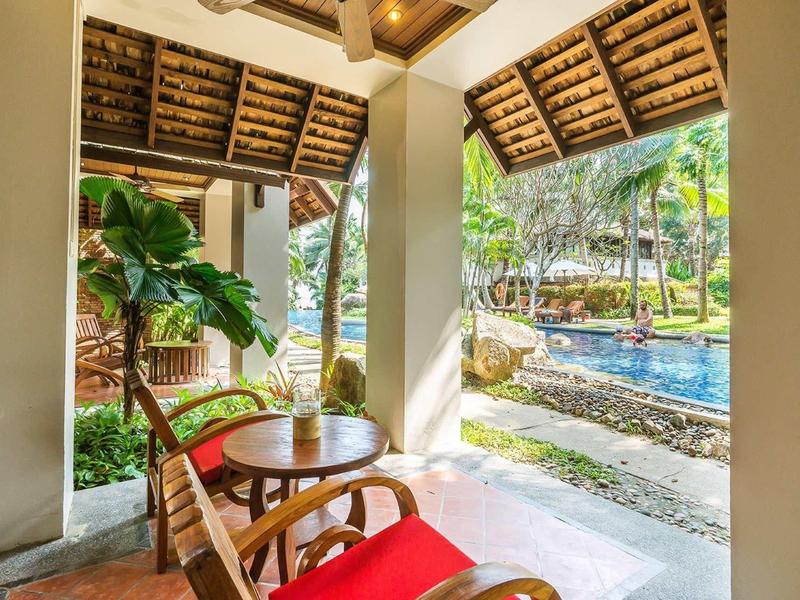 Pohodlný balkón s závěsným křeslem, dřevěným stolem, rostlinou a výhledem na bazén a zahradu.