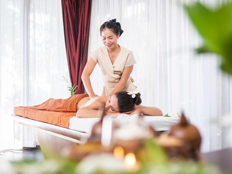 Žena dostává masáž zad v světlé wellness místnosti s bílými závěsy a červenými akcenty.