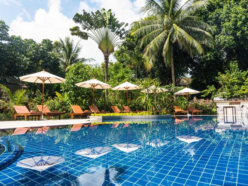 Duży niebieski basen z leżakami i parasolami otoczony tropikalną roślinnością.