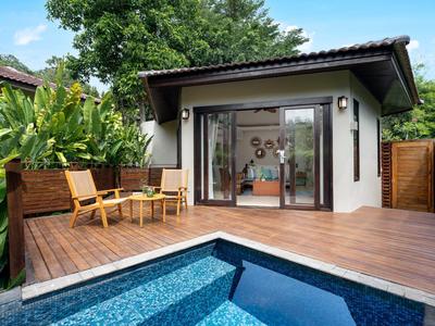 Bungalow moderne avec terrasse en bois, deux chaises et une petite piscine bleue dans un jardin tropical.