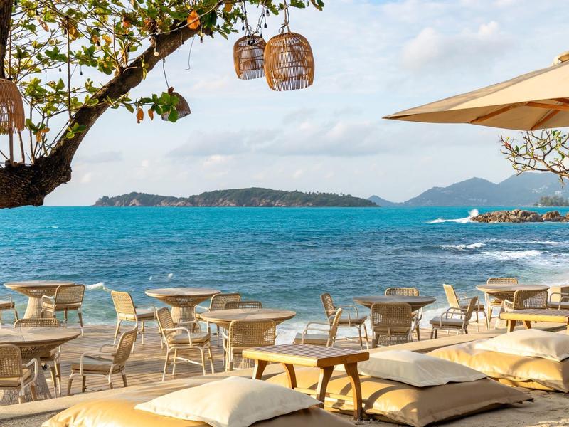 Espace extérieur confortable avec tables, chaises et coussins au bord de la mer sous des arbres et un parasol.