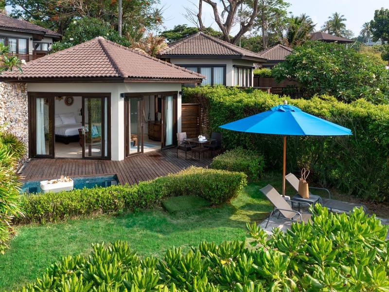 Une maison de vacances moderne avec terrasse, chaises de jardin et parasol bleu dans un cadre verdoyant.