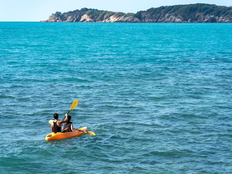 Deux personnes pagayant en kayak jaune sur une eau bleue calme avec une île en arrière-plan.