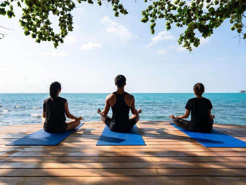 Trois personnes font du yoga sur un quai en bois face à la mer sous un ciel clair.