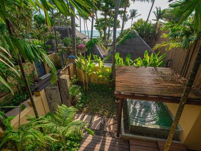 Tropischer Garten mit dichtem Grün, Bambushütte, Holzsteg und Blick auf ein Gebäude mit Terrasse.