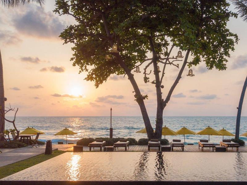 Tramonto sulla spiaggia con piscina, lettini, ombrelloni e palme.